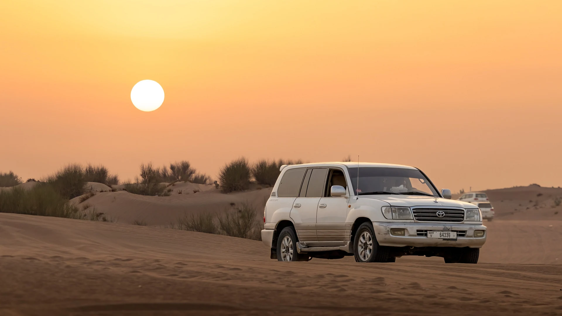 A white 4x4 parked on a desert dune outside Dubai at sunset, with the sun low over scrubby dune vegetation